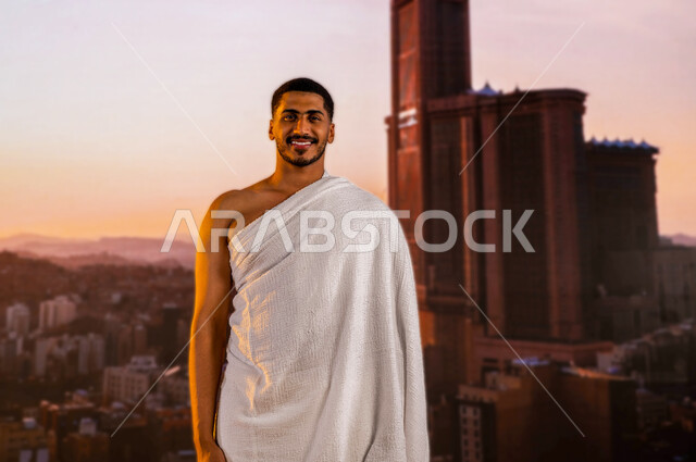 The background of the towers of the Great Mosque of Mecca and the clock tower, a smiling Saudi Arabian Gulf Muslim young man wearing the Ihram clothes, the arrival of the pilgrims of the interior to Mecca to perform the Hajj in the month of Dhul-Hijjah, t