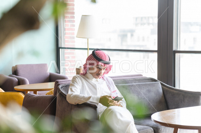 رجل عربي سعودي خليجي يحمل بيده هاتفه المحمول و يجلس في احد المطاعم  ، تصفح المواقع الالكترونية ، التسوق الالكتروني ،  كافيهات ومطاعم في السعودية 