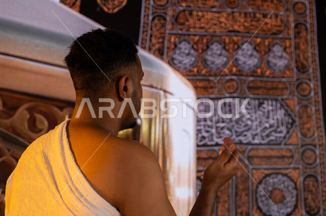 فضل العمرة في شهر رمضان المبارك، الدعاء والتقرب الى الله، صورة من الخلف لحاج اسمر البشرة عربي سعودي خليجي يرتدي ملابس الإحرام، يرفع يديه للأعلى ويدعو، الطواف حول الكعبة وآداء مناسك الحج والعمرة، خلفية الكعبة المشرفة ومقام إبراهيم عليه السلام