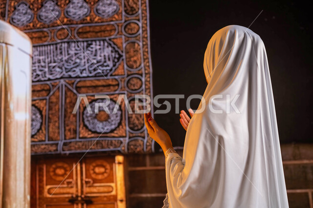 فضل العمرة في شهر رمضان المبارك، الدعاء والتضرع الى الله، صورة من الخلف لحاجة عربية سعودية خليجية ترتدي ملابس الصلاة البيضاء ترفع يديها و تدعو بخشوع, اداء مناسك الحج والعمرة, العبادة والتقرب الى الله، خلفية الكعبة المشرفة ومقام إبراهيم عليه السلام