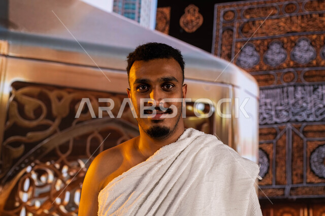 العبادة والتقرب الى الله، فضل العمرة في شهر رمضان المبارك، صورة مقربة لحاج اسمر البشرة مسلم عربي سعودي خليجي يرتدي ملابس الإحرام ينظر الى الكاميرا, آداء فريضة الحج والعمرة في شهر ذو الحجة، خلفية الكعبه المشرفه ومقام إبراهيم عليه السلام