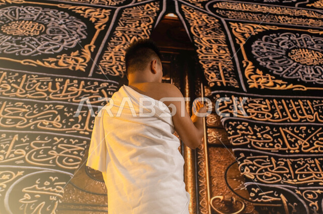 الدعاء والتضرع الى الله، صورة من الخلف لحاج عربي سعودي خليجي يرتدي ثياب الإحرام يرفع يديه و يدعو بخشوع, فضل العمرة في شهر رمضان المبارك، اداء مناسك الحج والعمرة, العبادة والتقرب الى الله، خلفية باب الكعبة المشرفة في الحرم المكي الشريف.
