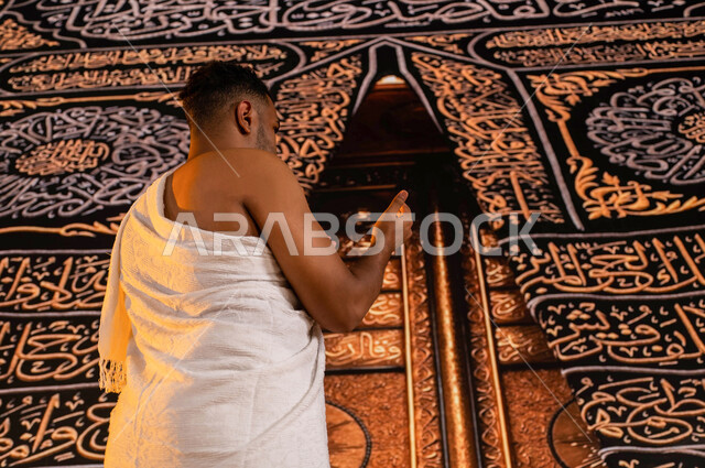فضل العمرة في شهر رمضان المبارك, الاجتهاد في الطاعات والتقرب إلى الله في الأشهر المباركة, صورة من الجانب لحاج عربي سعودي خليجي يرتدي ثياب الإحرام يرفع يديه و يدعي بخشوع، أداء فريضة الحج و العمرة في شهر ذو الحجة, خلفية باب الكعبة الذهبي في الحرم الشريف