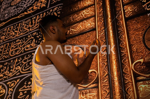 الدعاء والعباده والتقرب الى الله في مكة المكرمة، صورة من الجانب لشاب مسلم عربي سعودي خليجي يرفع يديه للأعلى ويدعي بخشوع، آداء مناسك الحج والعمرة، ارتداء ملابس الاحرام، موسم الحج 2023، الوقوف أمام الباب الذهبي للكعبة المشرفة في المسجد الحرام في مكة المكرمة