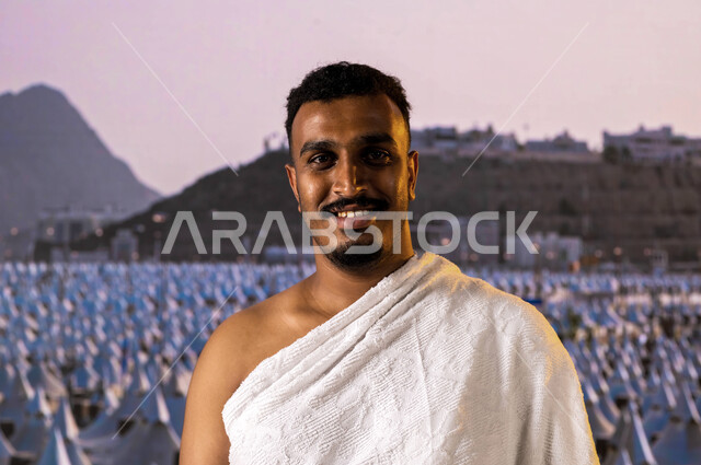 رجل أسمر مسلم خليجي عربي سعودي يبتسم للكاميرا ويرتدي ملابس الإحرام ،  أداء مناسك الحج والعمرة،  التقرب الى الله تعالى والتضرع له، المبيت في منى في موسم الحج 2023, قدوم الحجاج من داخل السعودية, خلفية مشعر منى والخيام البيضاء