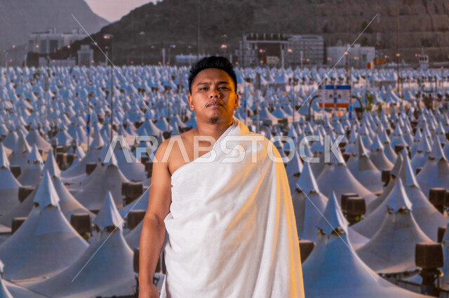 معالم دينية إسلامية، العبادة والتقرب إلى الله بأداء مناسك الحج والعمرة، حاج أندونيسي مسلم يرتدي ملابس الإحرام ينظر الى الكاميرا، المبيت في منى في موسم الحج 2023, قدوم الحجاج من خارج السعودية, خلفية مشعر منى والخيام البيضاء