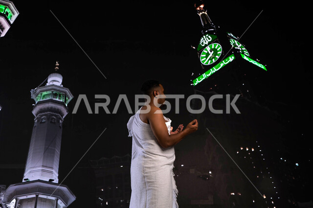 فضل العمرة في شهر رمضان المبارك، صورة من الخلف لحاج أسمر مسلم عربي خليجي سعودي يرتدي ملابس الإحرام، الدعاء والتوسل الى الله في مكة المكرمة، قدوم الحجاج من داخل السعودية, آداء مناسك الحج والعمرة، خلفية برج الساعة ومآذن الحرم المكي الشريف