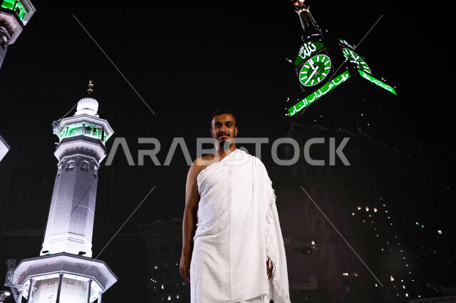 أداء العمرة في مكة المكرمة في شهر رمضان المبارك، رجل أسمر مسلم خليجي عربي سعودي يرتدي ملابس الإحرام ، أداء مناسك الحج والعمرة، قدوم الحجاج من داخل السعودية, الوقوف أمام المسجد الحرام في وقت الليل, خلفية برج الساعة ومآذن الحرم المكي الشريف