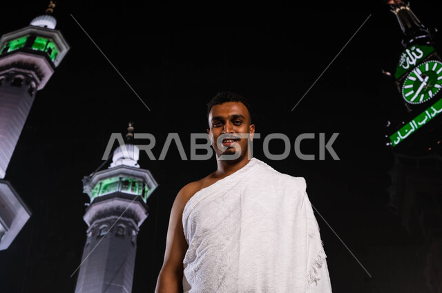 قدوم الحجاج من داخل السعودية, أداء العمرة في مكة المكرمة في شهر رمضان المبارك، رجل أسمر مسلم خليجي عربي سعودي يرتدي ملابس الإحرام ، أداء مناسك الحج والعمرة، الوقوف أمام المسجد الحرام في وقت الليل, خلفية برج الساعة ومآذن الحرم المكي الشريف