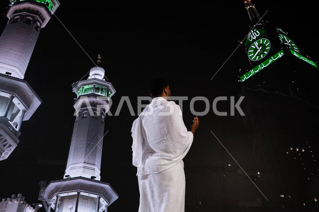 قدوم الحجاج من داخل السعودية, الدعاء والتقرب الى الله، فضل العمرة في شهر رمضان المبارك، صورة من الخلف لشاب مسلم عربي خليجي سعودي يرتدي ثياب الإحرام، الدعاء والتوسل الى الله في مكة المكرمة، آداء مناسك الحج والعمرة, خلفية برج الساعة ومآذن الحرم المكي الشريف