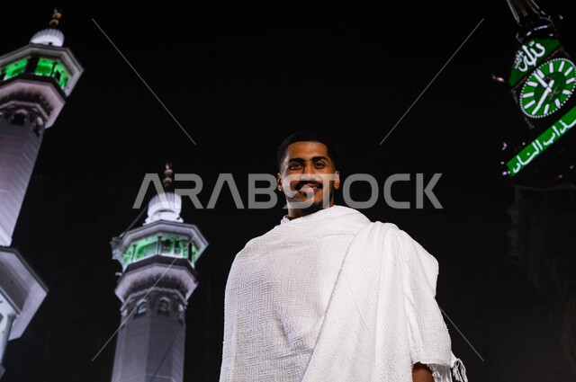 أداء العمرة في مكة المكرمة في شهر رمضان المبارك، رجل مسلم خليجي عربي سعودي يرتدي ملابس الإحرام، أداء مناسك الحج والعمرة، الوقوف أمام المسجد الحرام في وقت الليل, قدوم الحجاج من داخل السعودية, خلفية برج الساعة ومآذن الحرم المكي الشريف