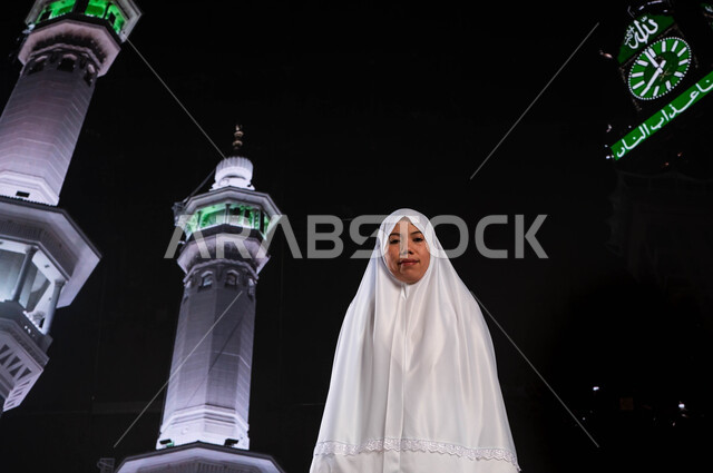 الوقوف أمام المسجد الحرام في وقت الليل, إمراة مسلمة أندونيسية ترتدي ملابس الاحرام تنظر إلى الكاميرا، التقرب الى الله بأداء الطاعات, قدوم الحجاج من خارج السعودية، آداء مناسك الحج والعمرة، خلفية برج الساعة ومآذن الحرم المكي الشريف 
