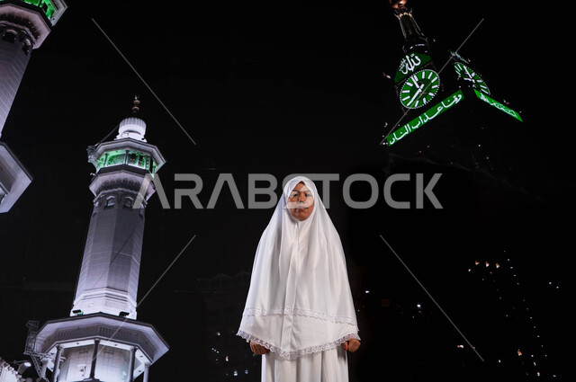 حاجة مسلمة أندونيسية ترتدي ملابس الاحرام، التقرب الى الله بأداء الطاعات, آداء مناسك الحج والعمرة، الوقوف أمام المسجد الحرام في وقت الليل, قدوم الحجاج من خارج السعودية, خلفية برج الساعة ومآذن الحرم المكي الشريف