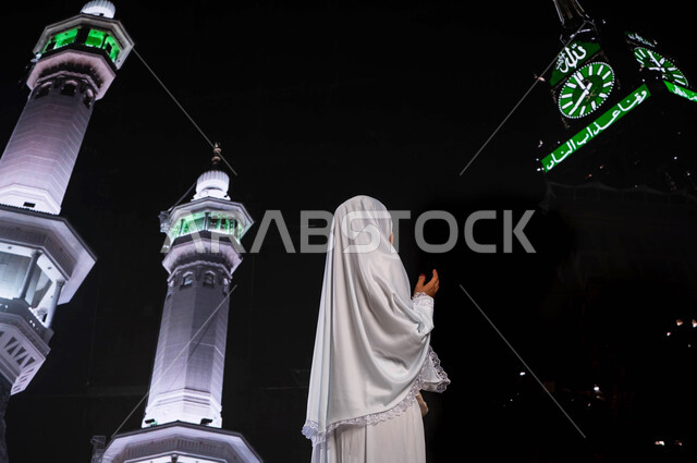 زيارة مكة المكرمة لأداء مناسك الحج والعمرة, صورة من الخلف لإمرأة مسلمة أندونيسية ترتدي ملابس الاحرام, الدعاء والتوسل الى الله في مكة المكرمة, التقرب الى الله تعالى وطاعته, قدوم الحجاج من خارج السعودية, خلفية خلفية برج الساعة ومآذن الحرم المكي الشريف