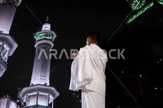 فضل العمرة في شهر رمضان المبارك، صورة من الخلف لشاب أندونيسي مسلم يرتدي ملابس الإحرام، الدعاء والتوسل الى الله في مكة المكرمة، آداء مناسك الحج والعمرة، الحرم المكي الشريف, خلفية برج الساعة ومآذن الحرم المكي الشريف