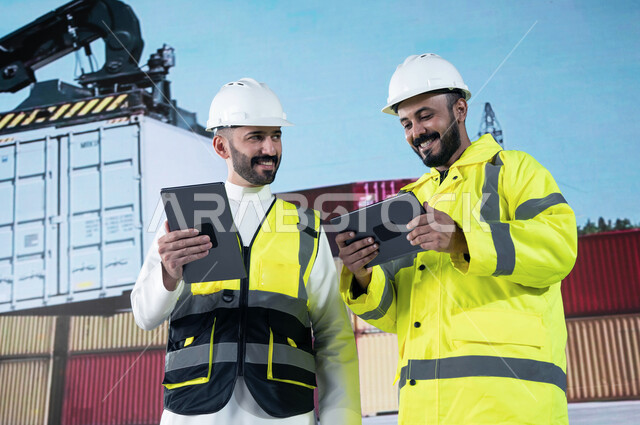 مراجعة قوائم الشحن البري عبر أجهزة التابلت, ميناء شحن البضائع و الحاويات التجاريه، صورة مقربة لمهندسان عربيان سعوديان خليجيان مبتسمان يقومان بالاشراف على توصيل ونقل الكونتينرات، استخدام الآلات المتطوره الحديثه في الإستيراد والتصدير.