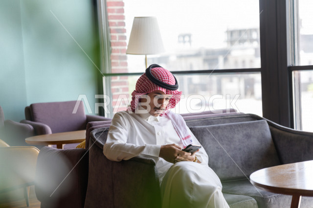 رجل عربي سعودي خليجي يحمل بيده هاتفه المحمول و يجلس في احد المطاعم  ، تصفح المواقع الالكترونية ، التسوق الالكتروني ،  كافيهات ومطاعم في السعودية 