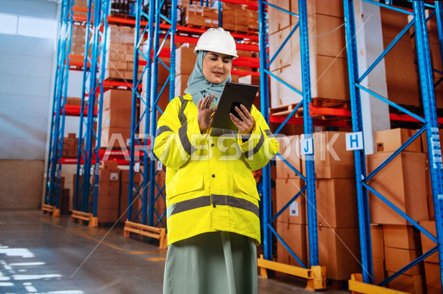 التأكد من وجود البضائع, عمليات التصنيف والترتيب والترميز للمواد المطلوبة, تحديد مستوى المخزون في المستودع، صورة مقربة لمهندسة عربية خليجية سعودية في المخزن تحمل بيدها جهاز التابلت المحمول, خلفية صناديق وكراتين مغلفة.