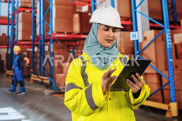 خلفية صناديق وكراتين مغلفة, عمليات التصنيف والترتيب والترميز للمواد المطلوبة, تحديد مستوى المخزون في المستودع، صورة مقربة لمهندسة عربية خليجية سعودية في المخزن تحمل بيدها جهاز التابلت المحمول للتأكد من توفر البضاعة.