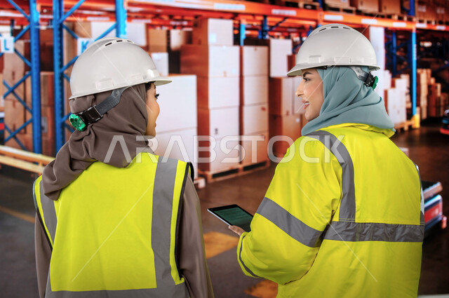 Determining the stock level in the warehouse, sorting, arranging and coding the required materials, use the tablets to ensure the availability of the goods, a close-up of the back of two Saudi Arab Gulf engineers in the warehouse, the background of boxes 