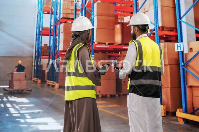 عمليات مطابقة المشتريات مع المواصفات المطلوبة, تحضير طلبيات جديدة, تصوير من الخلف لمدير ترتيب وتنظيم المستودعات المحلية ومهندسة عربية سعودية خليجية يستخدمان أجهزة التابلت, دمج العمل مع التقنية و التكنولوجيا, جرد البضائع في المستودع، خلفية كراتين مغلفة.