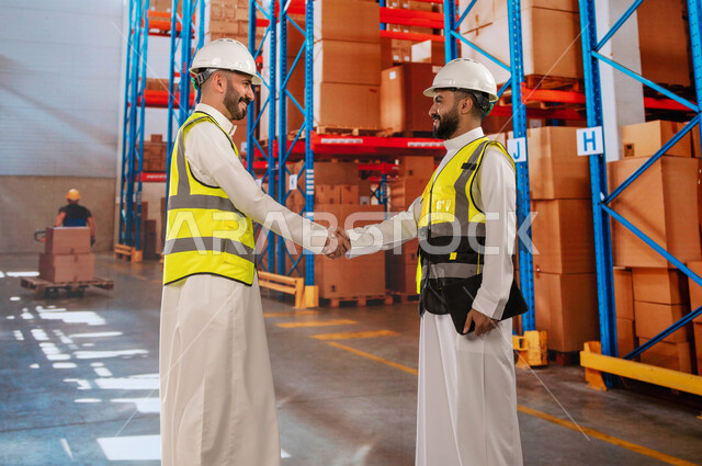 التصافح لإنجاز و نجاح خطة العمل المتفقة, دمج العمل مع التقنية و التكنولوجيا, منتجات و بضائع عربية سعودية خليجية، عمليات جرد و مطابقة البضائع في المستودع، صورة من الخلف لمهندسان عربيان خليجيان سعوديان في المخزن، خلفية صناديق وكراتين مغلفة.