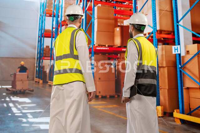 منتجات و بضائع عربية سعودية خليجية، دمج التقنية و التكنولوجيا مع العمل, عمليات جرد و مطابقة البضائع في المستودع، تصوير من الخلف لمدير ترتيب وتنظيم المستودعات المحلية ومهندس عربي سعودي خليجي يحمل بيده جهاز التابلت، خلفية صناديق وكراتين مغلفة.