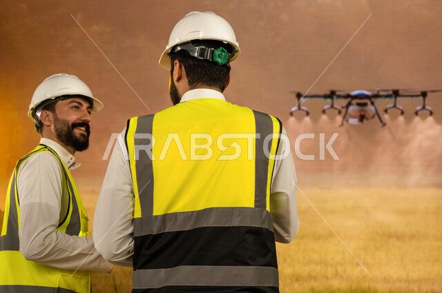 زيادة انتاج وجودة المحاصيل الزراعيه, تطور التقنيه والتكنولوجيا الحديثه في الري وسقي المزروعات, درون لسقي الاراضي الزراعيه في السعوديه,     صورة من الخلف لمهندسان زراعيان عربيان سعوديان خليجيان يرتديان خوذة وسترة الحمايه,  آلات واجهزه متطوره