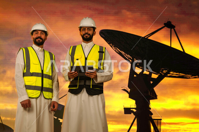 تتبع حركة الكواكب والنجوم عبر الستلايت، متابعة اتصال القمر الصناعي مع الفضاء الخارجي، مهندسان عربيان خليجيان سعوديان يرتديان الخوذة وسترة الحماية الخاصة, إستخدم جهاز التابلت للإشراف على عمل الأقمار الصناعية، علم الفلك والفضاء، خلفية غروب الشمس. 