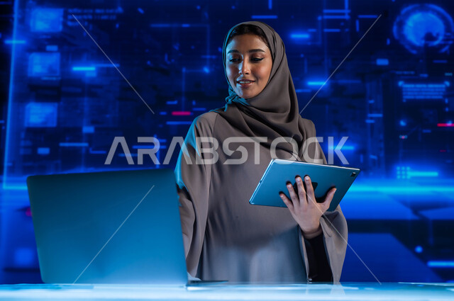 تطوير الشركات والمشاريع الاقتصادية، سيدة أعمال عربية خليجية سعودية محجبة في مقر عملها بالشركة تقوم بمتابعة البورصة من خلال الحاسوب المحمول والتابلت، الاستثمار المالي والتجارة الالكترونية، خلفية هولوجرامية ثلاثية الأبعاد.