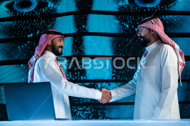صفقات عمل في شركات التقنية والتكنولوجيا المستقبلية الحديثة, رجلا أعمال عربيان سعوديان خليجيان يرتديان الزي السعودي التقليدي يقومان بالمصافحة, خلفية شاشات هولوجرامية ثلاثية الأبعاد للذكاء الاصطناعي، رؤية مستقبيلة ٢٠٣٠.