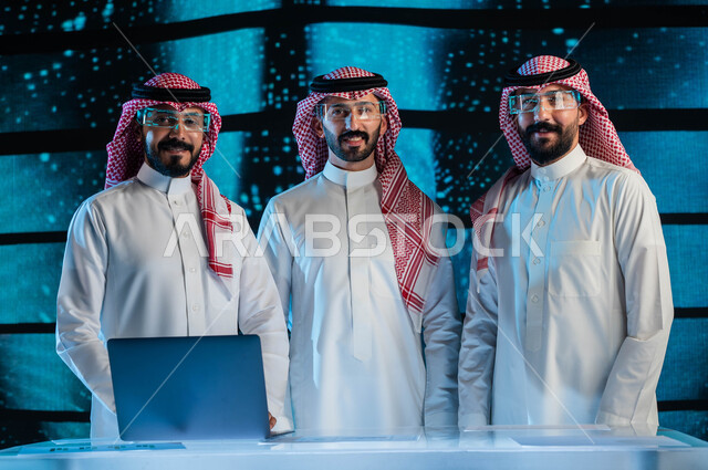  النظر إلى الكاميرا، صفقات عمل في شركات التقنية المستقبلية الحديثة, مجموعة من رجال أعمال عرب سعوديين خليجيين يرتدون الزي السعودي التقليدي ونظارات الواقع الافتراضي, خلفية شاشات هولوجرامية ثلاثية الأبعاد للذكاء الاصطناعي، رؤية مستقبيلة ٢٠٣٠.