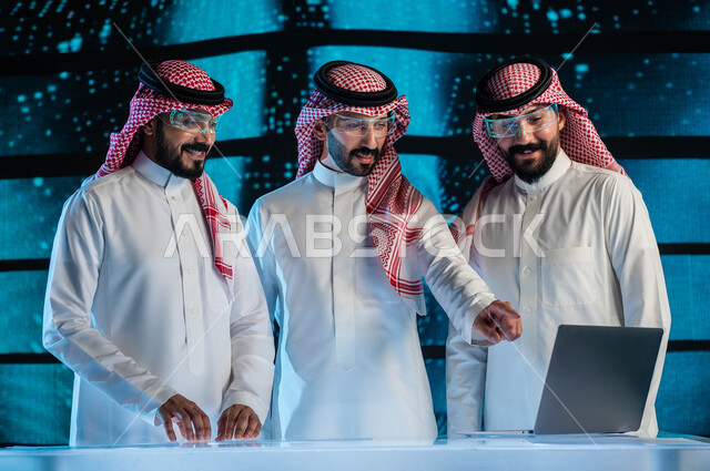 إجراء صفقات عمل في شركات التقنية والتكنولوجيا المستقبلية الحديثة, مجموعة من رجال أعمال عرب سعوديين خليجيين يرتدون الزي السعودي التقليدي ونظارات الواقع الافتراضي, خلفية شاشات هولوجرامية ثلاثية الأبعاد للذكاء الاصطناعي، رؤية مستقبيلة ٢٠٣٠.