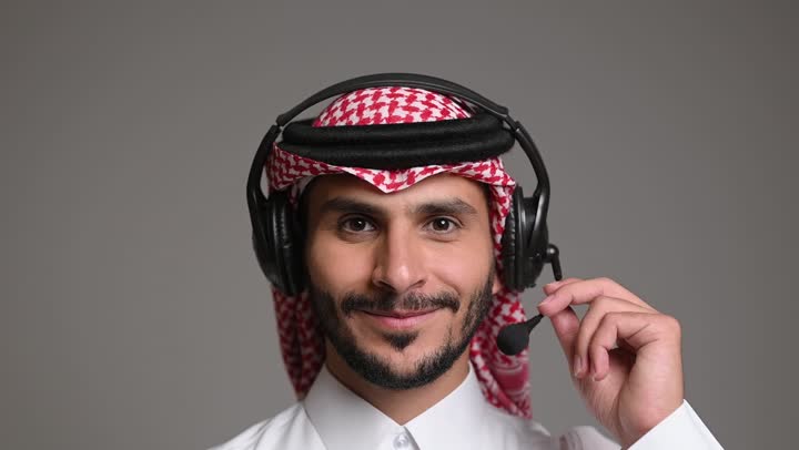 Answering and responding to inquiries and queries, communicating with customers to solve problems and complaints, using advanced technologies in work, close-up portrait of a Saudi Arabian Gulf employee wearing a traditional thobe and shemagh adjusting a headset, working in the customer service department, gray background