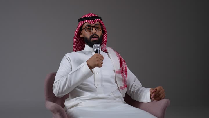 Effective communication, exchange of dialogues and discussions, conducting interviews and asking questions, a young Saudi Arabian Gulf man wearing traditional thobe and shemagh sitting on a chair holding a loudspeaker and giving speeches, holding workshops and training courses, gray background