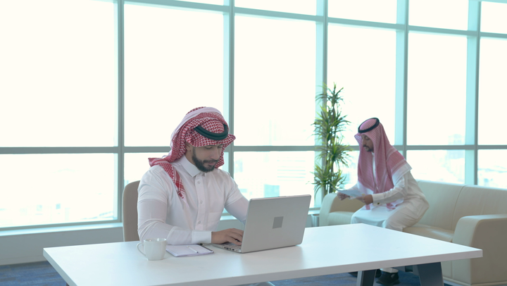 A Saudi Arabian Gulf employee using a laptop, discussing work with a colleague in the company, distributing the company’s tasks and work, the Saudi Gulf work environment, guidance and direction