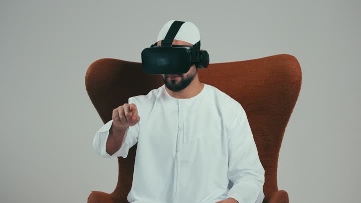 Clicking and merging and interacting, trying to touch something, side portrait of an Arab Gulf Emirati man wearing a hat, kandura and virtual reality glasses sitting on a chair raising his hands and simulating the imaginary world, using modern advanced technology and technology, white background