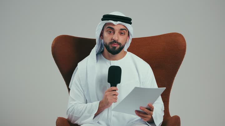 Effective communication, dialogue and discussion, conducting interviews and asking questions, portrait of an Emirati Gulf Arab man wearing a kandura and ghutra sitting on a chair holding a loudspeaker and a white paper, giving speeches and looking at the camera, gray background