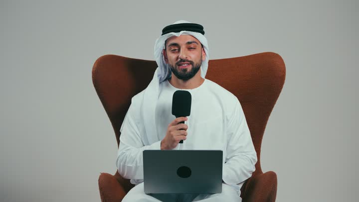 اجراء المقابلات وطرح الأسئلة، استخدام الأجهزة التقنية الحديثة، بورتريه لرجل عربي خليجي اماراتي يرتدي الكندوره والغتره البيضاء يجلس على الكرسي يستخدم الحاسوب المحمول ويحمل بيده مكبر الصوت يقوم بإلقاء الخطابات، خلفية رمادية