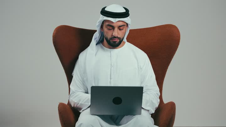 اجراء مقابلة عمل عن بعد, انجاز الاعمال والمهام عن بعد عبر الحاسوب المحمول، تصفح الانترنت ووسائل التواصل الاجتماعي، بورتريه لرجل عربي اماراتي مبتسم يرتدي الكندوره والغتره البيضاء يجلس على كرسي مريح يقوم باستخدام الحاسب، خلفية رماديه