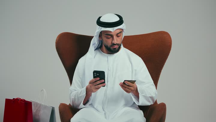 Electronic payment, bank credit cards, portrait of a young Emirati Gulf Arab man wearing a white kandura and ghutra sitting on a comfortable chair with colorful paper bags next to him, conducting a banking transaction via payment applications, facilitating remote payment, gray background