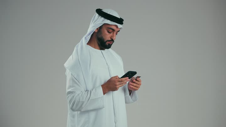 إمكانية تسديد الفواتير عن طريق البطافات الالكترونية، استخدام الوسائل والتقنيات الحديثة المتطورة، بورتريه من الجانب لرجل عربي خليجي اماراتي يرتدي الكندورة والغترة البيضاء يقوم بالدفع باستخدام بطاقة الإئتمان البنكية، خلفية رماديه