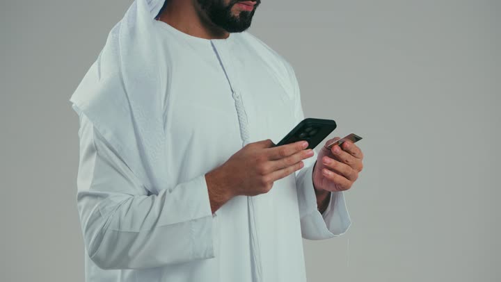 The ability to pay bills using electronic cards, using modern advanced means and technologies, a side portrait of an Emirati Gulf Arab man wearing a white kandura and ghutra, paying using a bank credit card, gray background