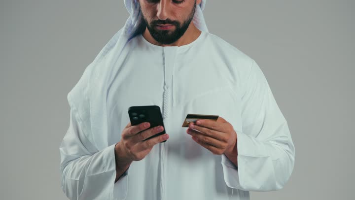 الدفع الإلكتروني عبر التقنيات الحديثة المتطورة، استخدام تطبيقات الجوال للدفع والشراء، بورتريه لشاب عربي خليجي اماراتي يرتدي الكندوره والغتره يمسك بيده البطاقة الإلكترونية والهاتف المحمول ينظر بإيماءات البشاشة والسرور، خلفية رماديه