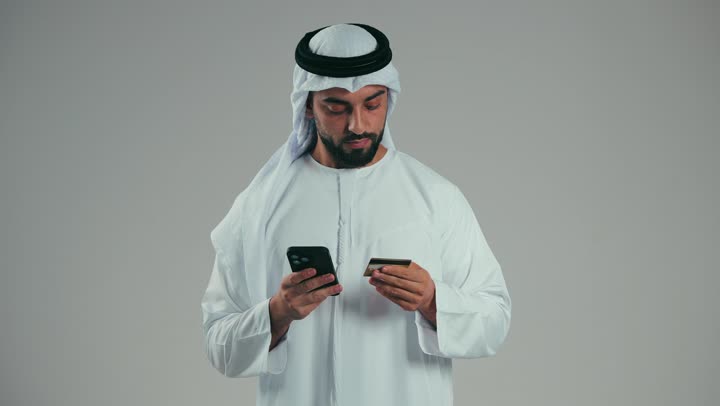 الدفع الإلكتروني عبر التقنيات الحديثة المتطورة، استخدام تطبيقات الجوال للدفع والشراء، بورتريه لشاب عربي خليجي اماراتي يرتدي الكندوره والغتره يمسك بيده البطاقة الإلكترونية والهاتف المحمول ينظر بإيماءات البشاشة والسرور، خلفية رماديه