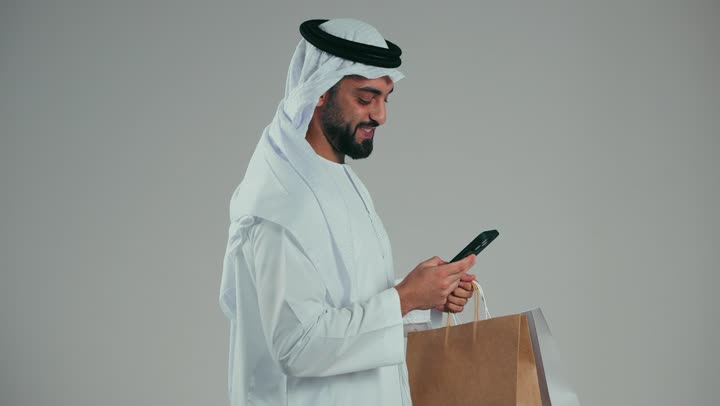 رجل عربي خليجي اماراتي يرتدي الكندوره والغتر البيضاء يستعمل البطاقه البنكيه, التسوق الالكتروني, طلبات اونلاين, الدفع عن طريق بطاقة الإئتمان, الشراء عبر برامج وتطبيقات الهاتف المحمول, خلفيه رماديه