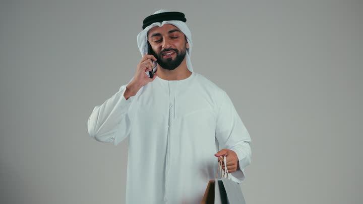 تقديم الهدايا لشخص ما، بورتريه لشاب عربي خليجي اماراتي مبتسم يرتدي الكندوره والغتره البيضاء يجري مكالمه هاتفيه يحمل اكياس ورقيه ملونه، معامله بنكيه عبر تطبيقات الدفع الالكتروني، تسهيل الدفع عن بعد، ايماءات السرور والسعاده بالتسوق، خلفيه رماديه