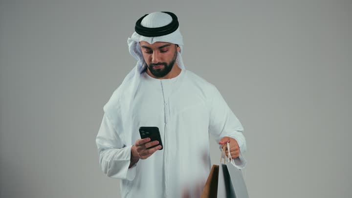 رجل عربي خليجي اماراتي يرتدي الكندوره والغتر البيضاء يستعمل البطاقه البنكيه, التسوق الالكتروني, طلبات اونلاين, الدفع عن طريق بطاقة الإئتمان, الشراء عبر برامج وتطبيقات الهاتف المحمول, خلفيه رماديه