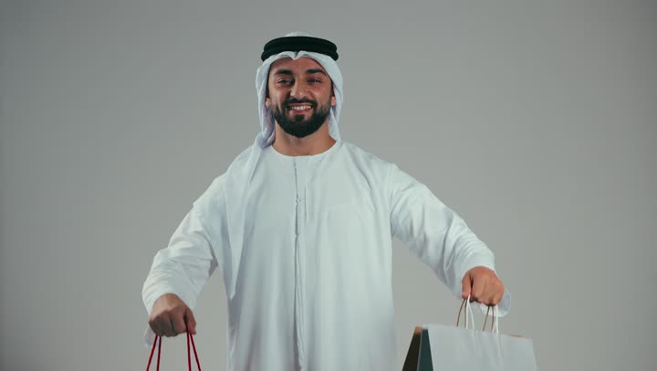 Portrait of an Emirati Gulf man wearing a white kandura and ghutra carrying colorful shopping bags, buying gifts, entertainment and shopping, gray background