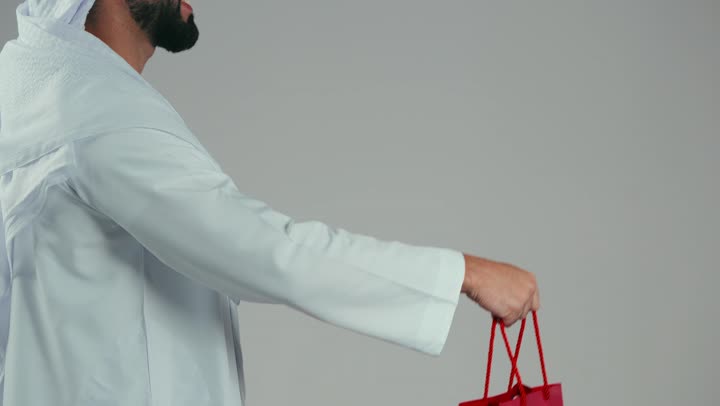 التسوق وشراء المستلزمات والهدايا عبر الانترنت، استخدام التكنولوجيا في الحياة اليومية، ايماءات وتعابير السرور، بورتريه من الجانب لشاب عربي خليجي اماراتي يرتدي الغترة والكندورة يمسك بيده الهاتف المحمول وأكياس التسوق، خلفية بيضاء